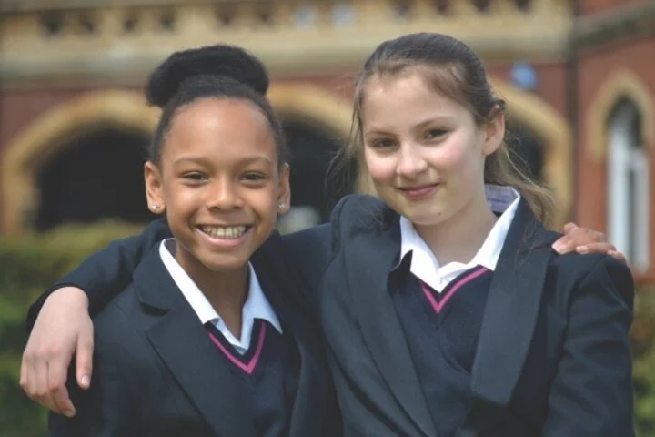 Two young students in school uniforms smiling and embracing outdoors, in front of their girls' boarding school in Surrey (11+ and 13+ entry).