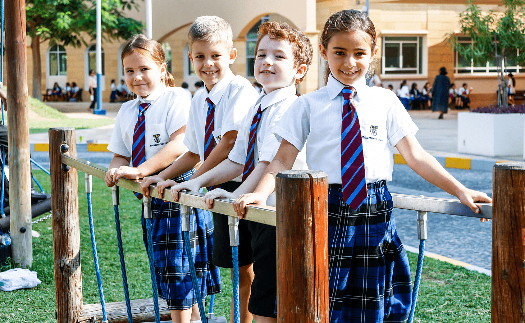 Four children in school uniforms standing on a playground, holding onto a horizontal bar, with a school building and other students in the background.