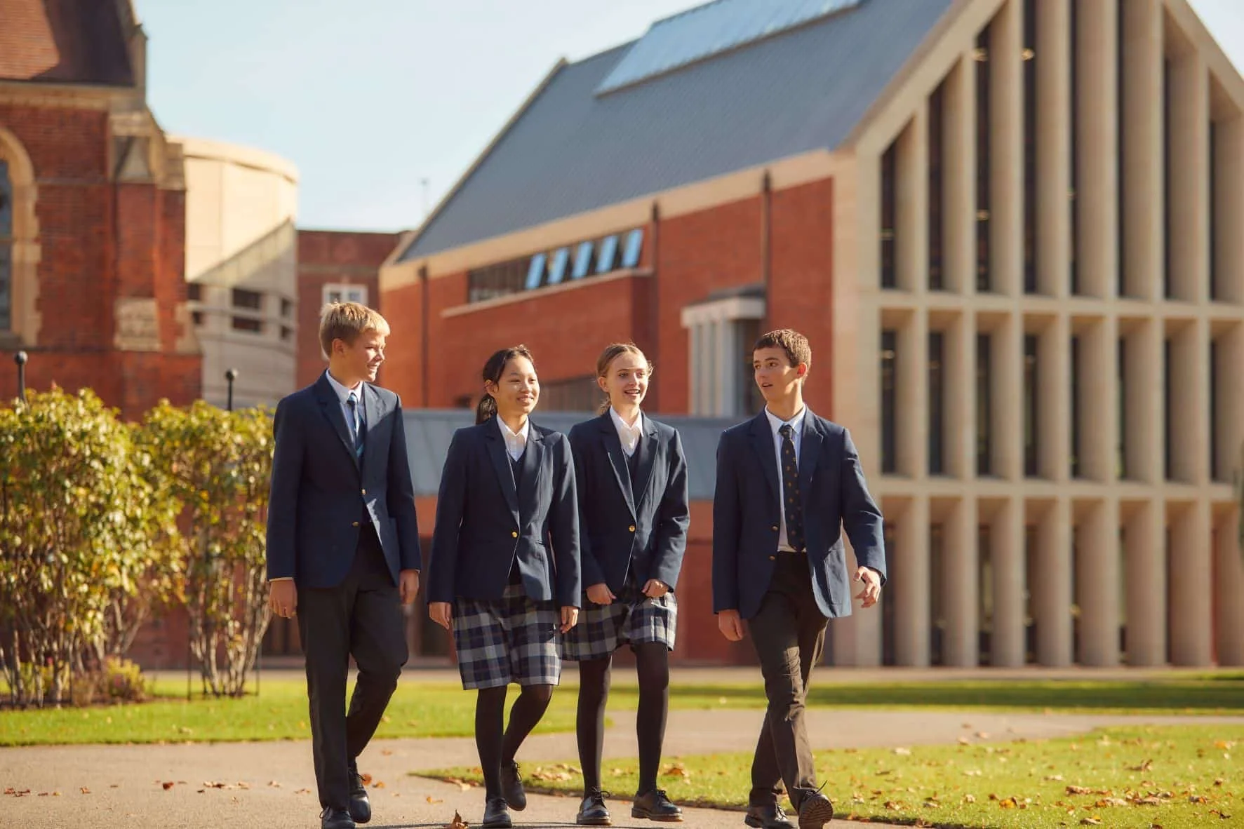 Four co-ed students in school uniforms walking on a 13+ entry boarding school campus with modern brick buildings in the background.