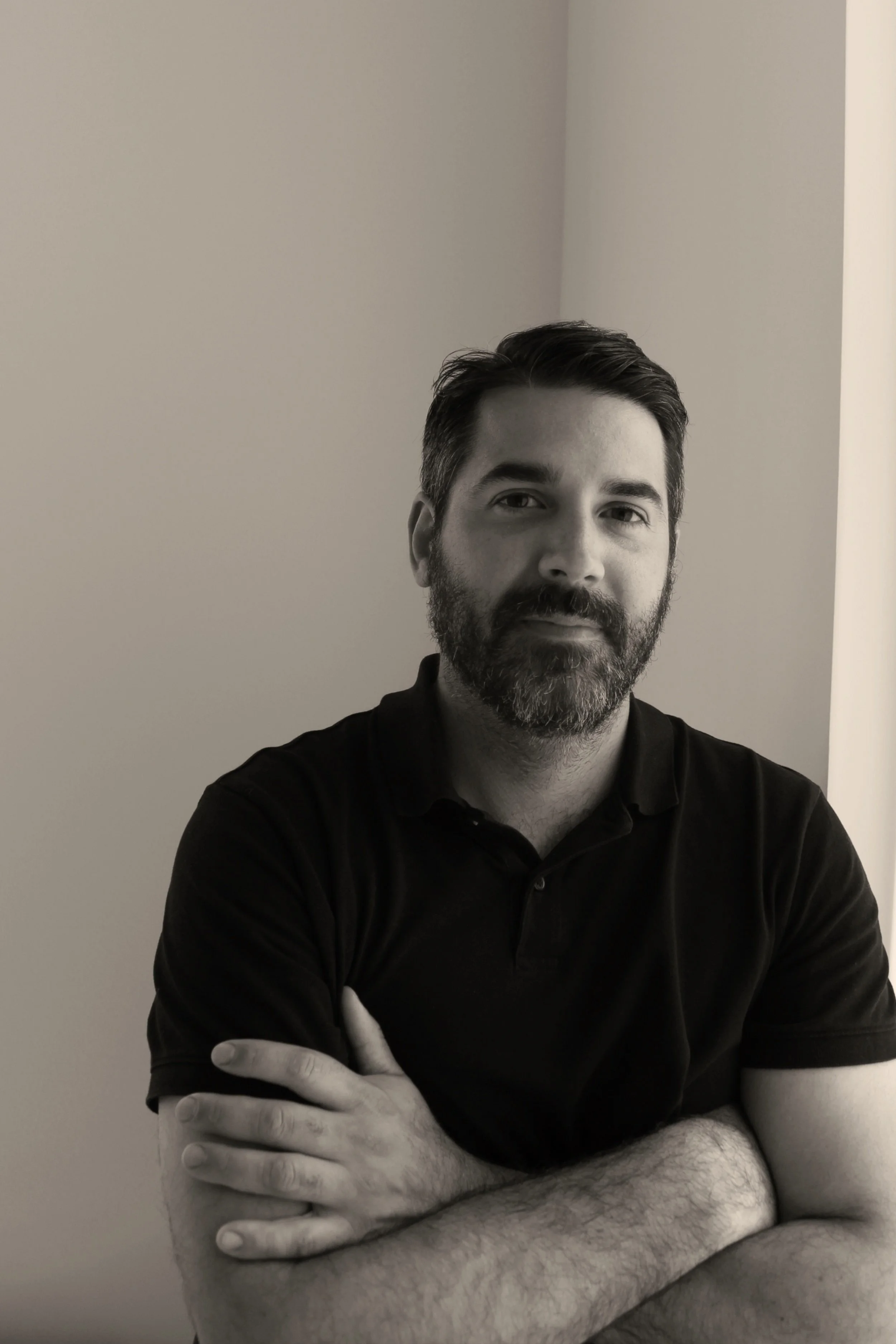 Black and white portrait of a man with dark hair and a beard, wearing a black t-shirt, standing against a plain wall.
