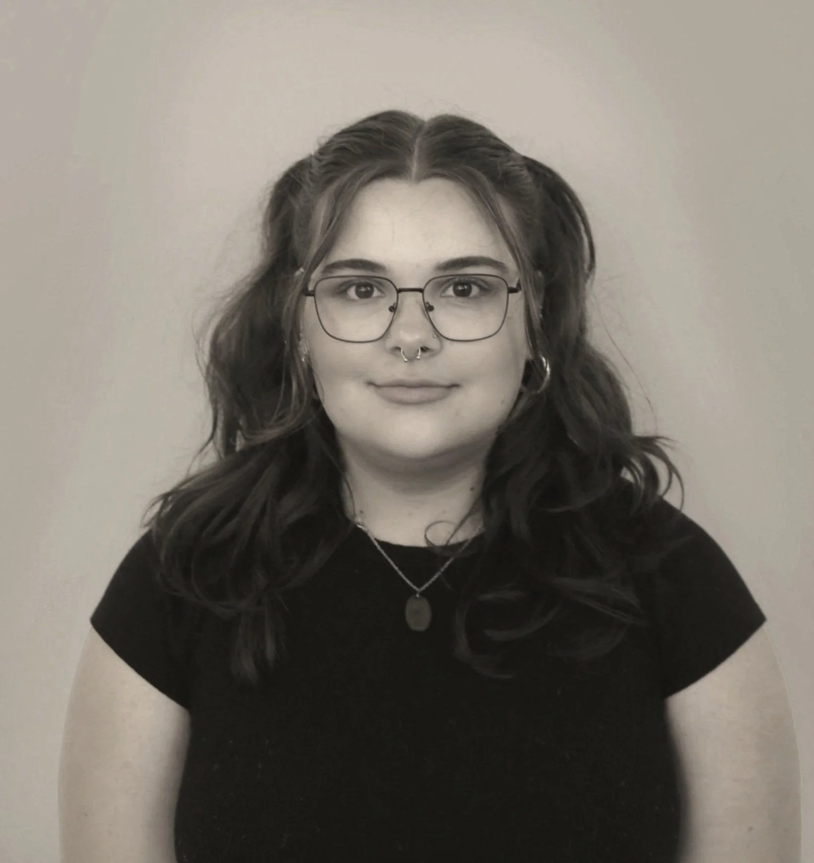 A young woman with wavy hair, glasses, and a septum piercing, wearing a black shirt and a necklace, smiling at the camera against a plain background.