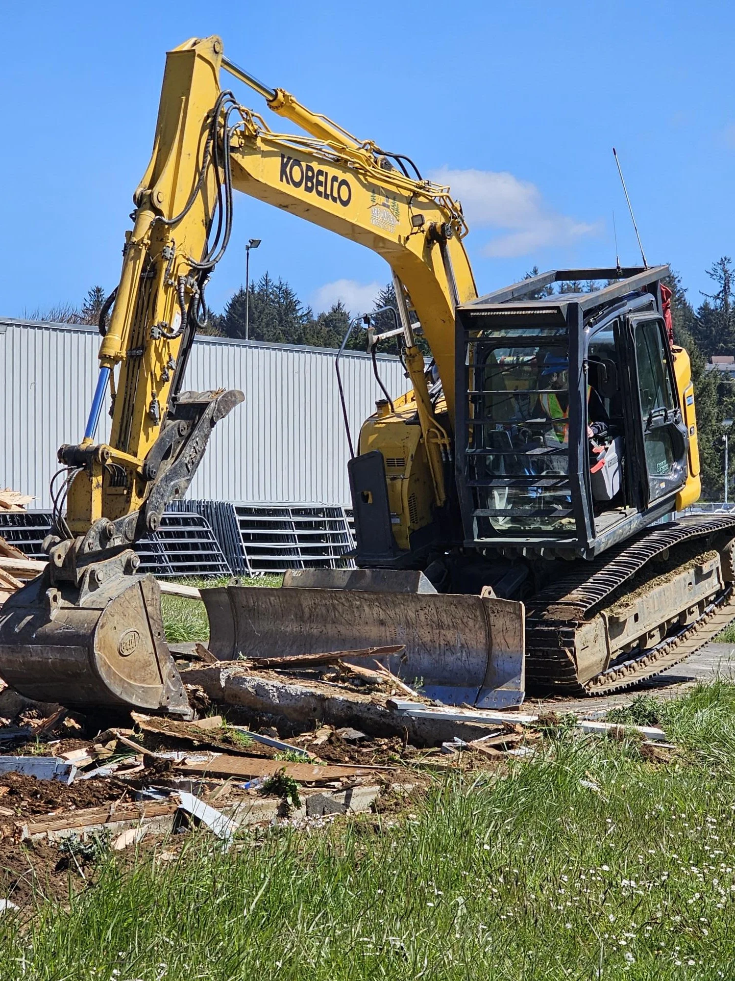 A bulldozer working on a dirt road in a forested area with tall trees and a clear sky.