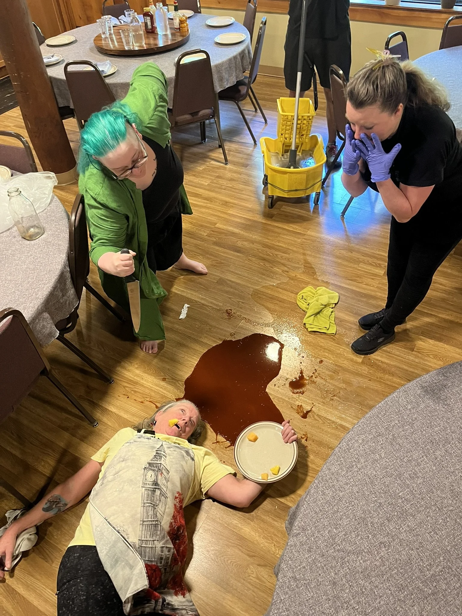 A woman lying next to a splatter of spilled sauce so that it looks like blood, while a Renegade member stands over her with a knife and a staffer pretends to be shocked. In the background is another staffer with the mop bucket looking incredibly bore