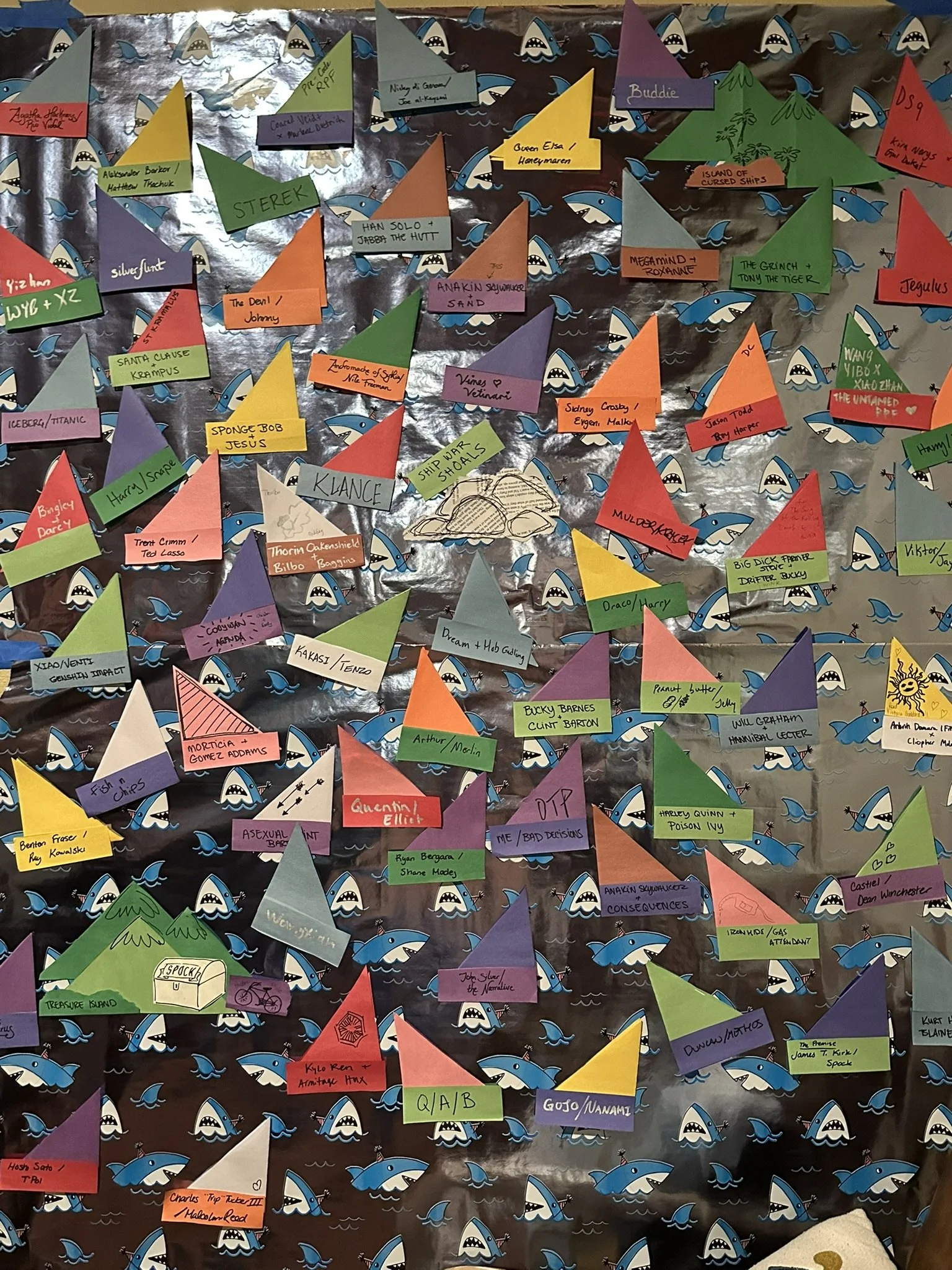 Shark wrapping paper with a bunch of colorful paper boats on it where people have written their favorite ships such as Klance, The Grinch/Tony The Tiger, and Anakin/Consequences