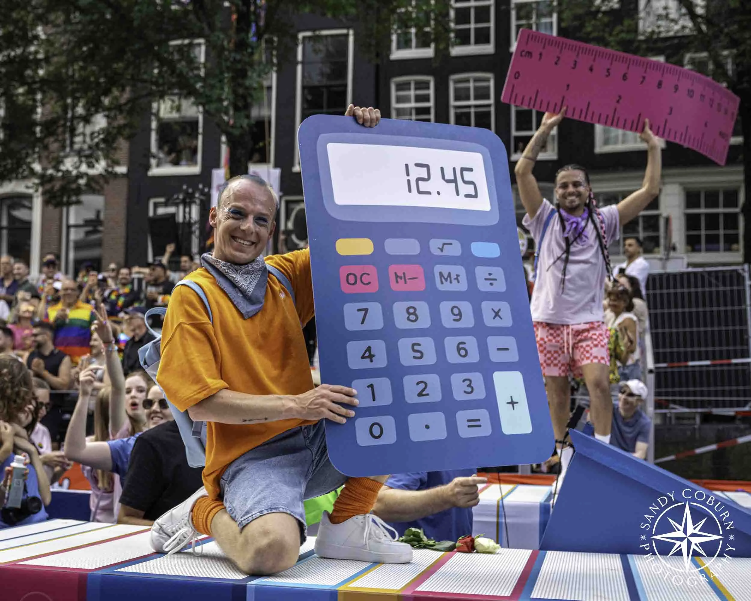 A participant poses with a calculator on the Ministry of Culture, Education, and Science (OCW) boat at the 2026 WorldPride Amsterdam Canal Parade