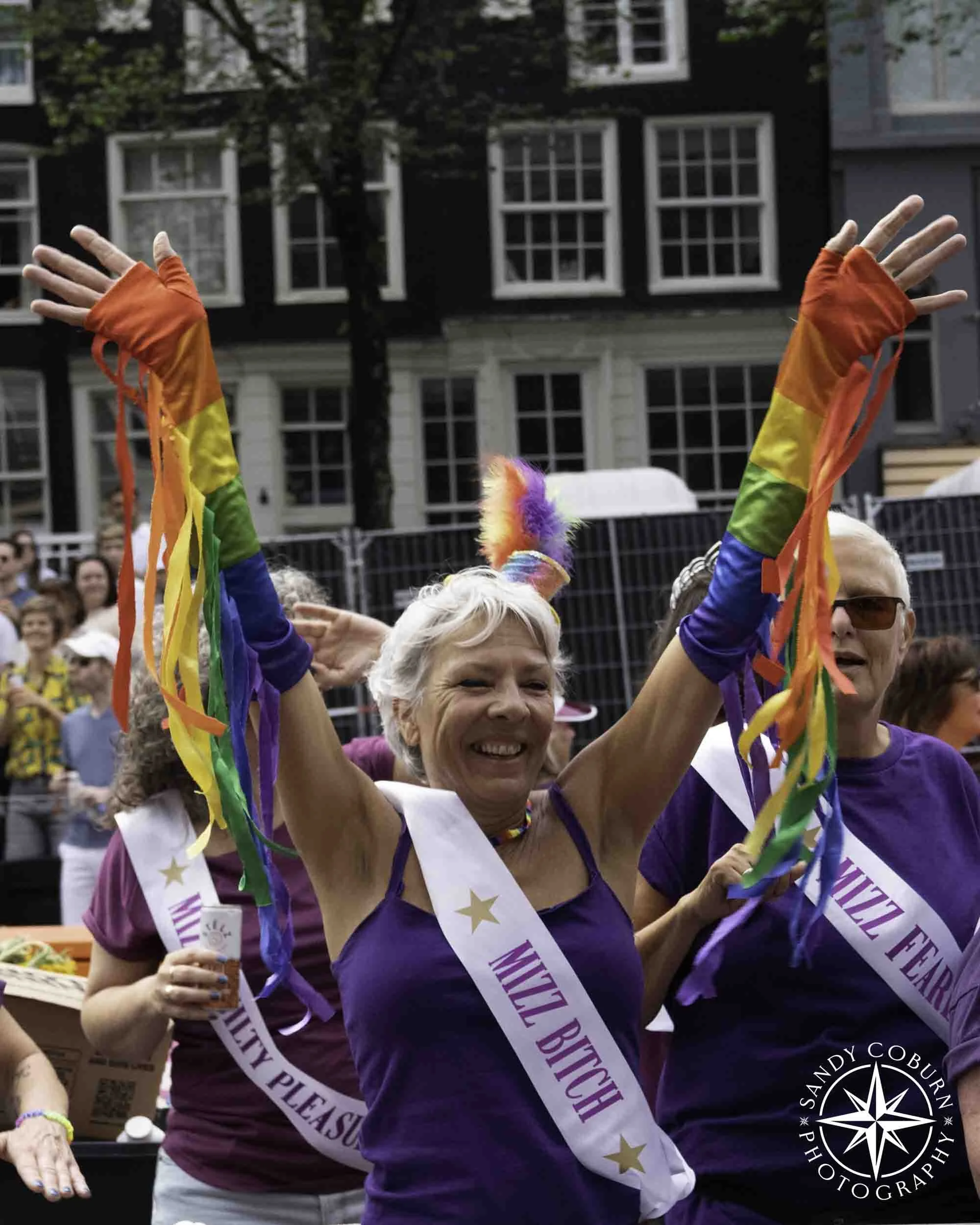 A person wearing rainbow pride fringed gloves and a sash labeled Miz Bitch waves to the crowd at the 2026 WorldPride Amsterdam Canal Parade