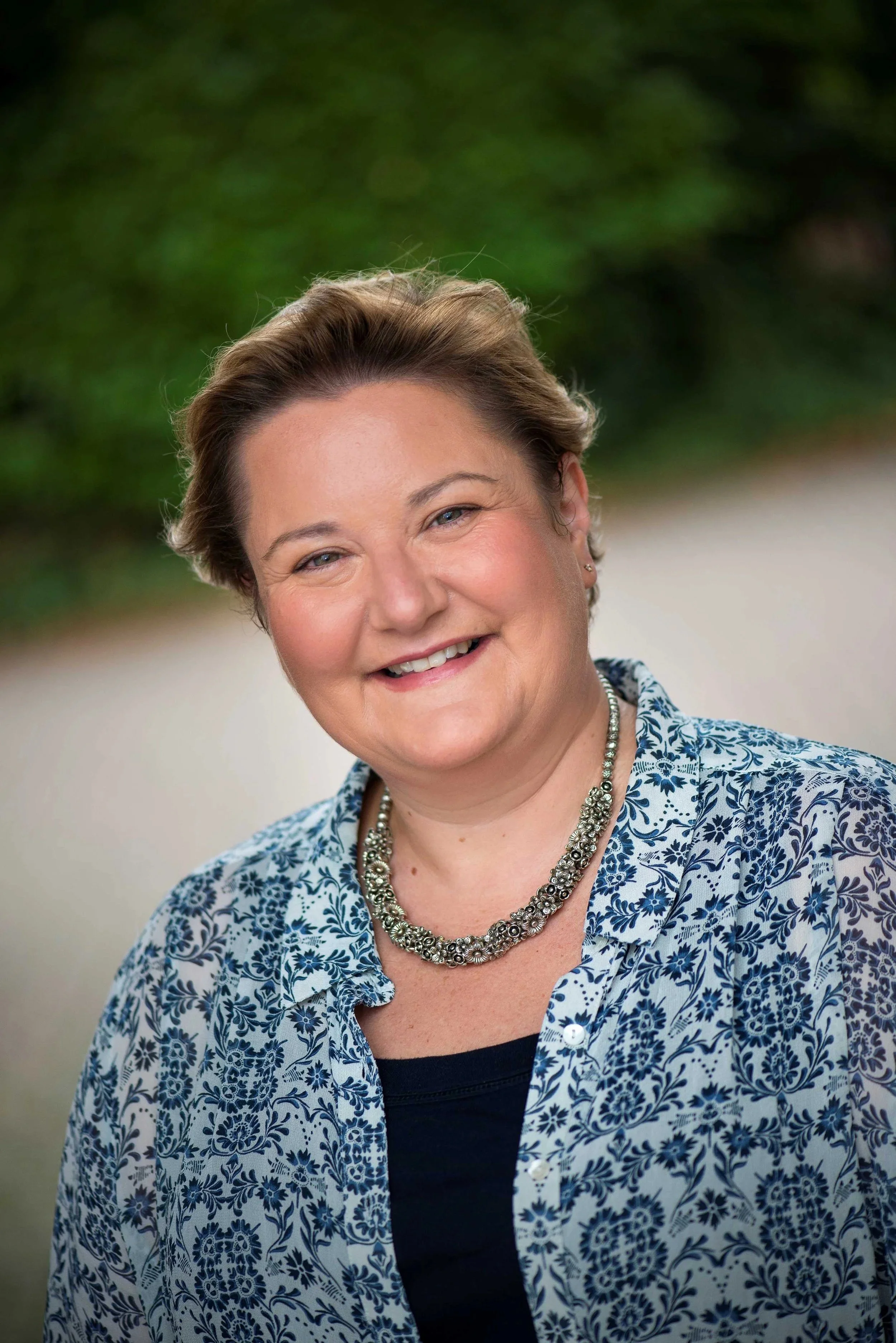 A smiling woman with short brown hair, wearing a patterned blue and white shirt and a floral necklace, standing outdoors with blurred green foliage in the background.