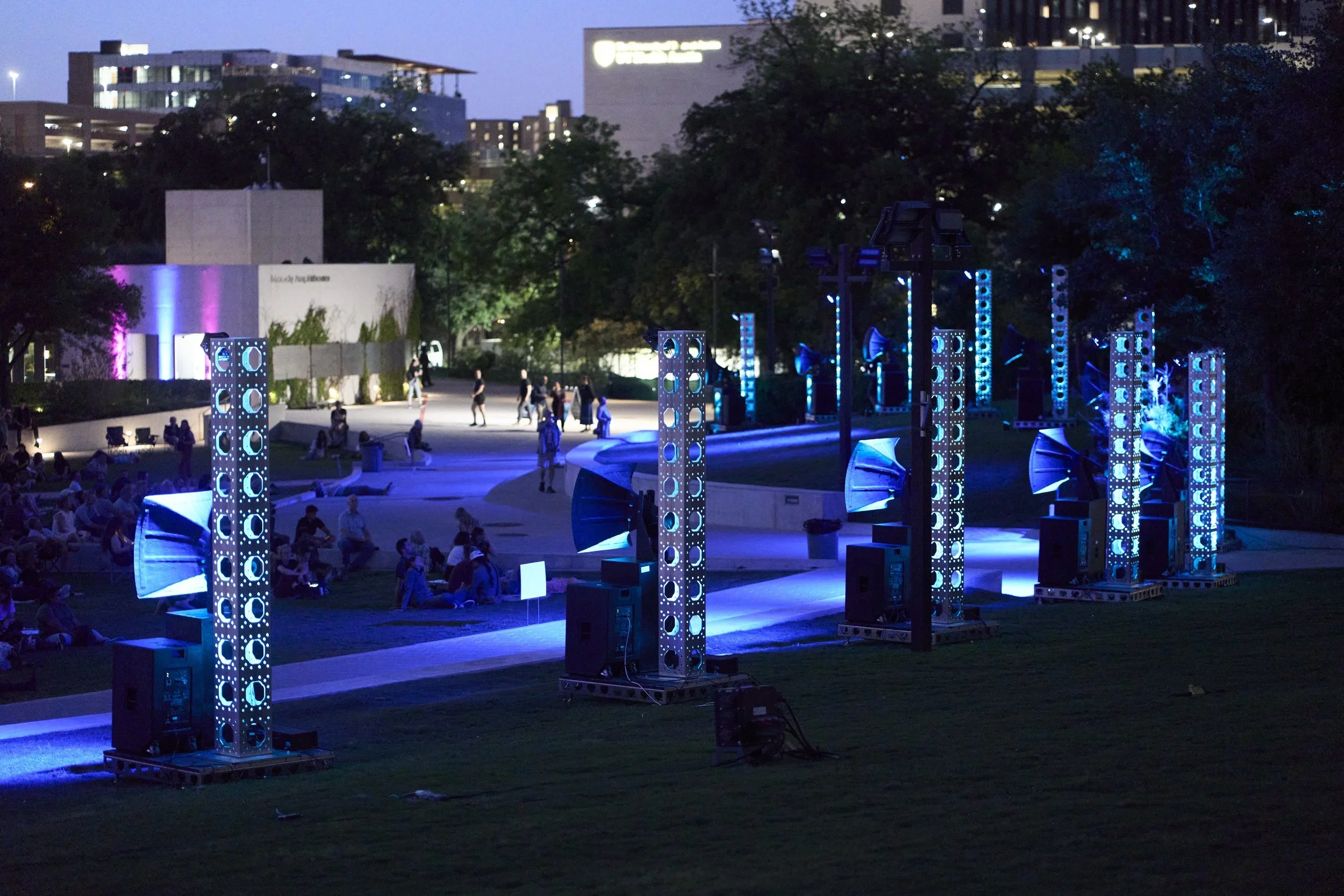 An outdoor evening scene with people gathering on a park lawn. There are illuminated blue and purple light structures, and in the background, city buildings with lights, including a large building with an illuminated sign.