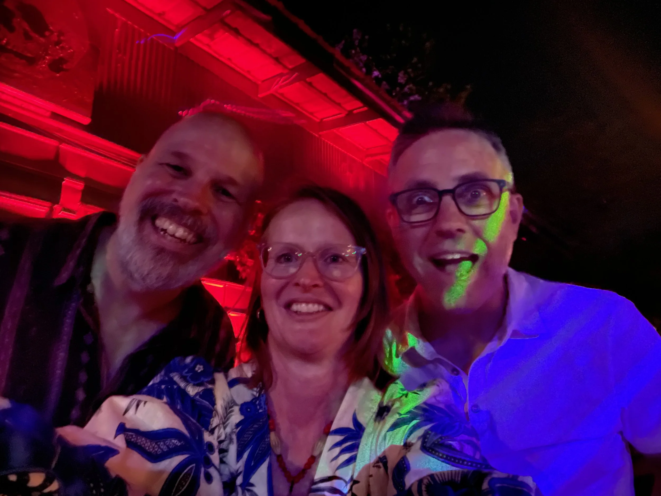 A group of people smiling in a setting with colorful and vibrant lighting, including red, green, and purple lights, likely at a nightlife venue or party.
