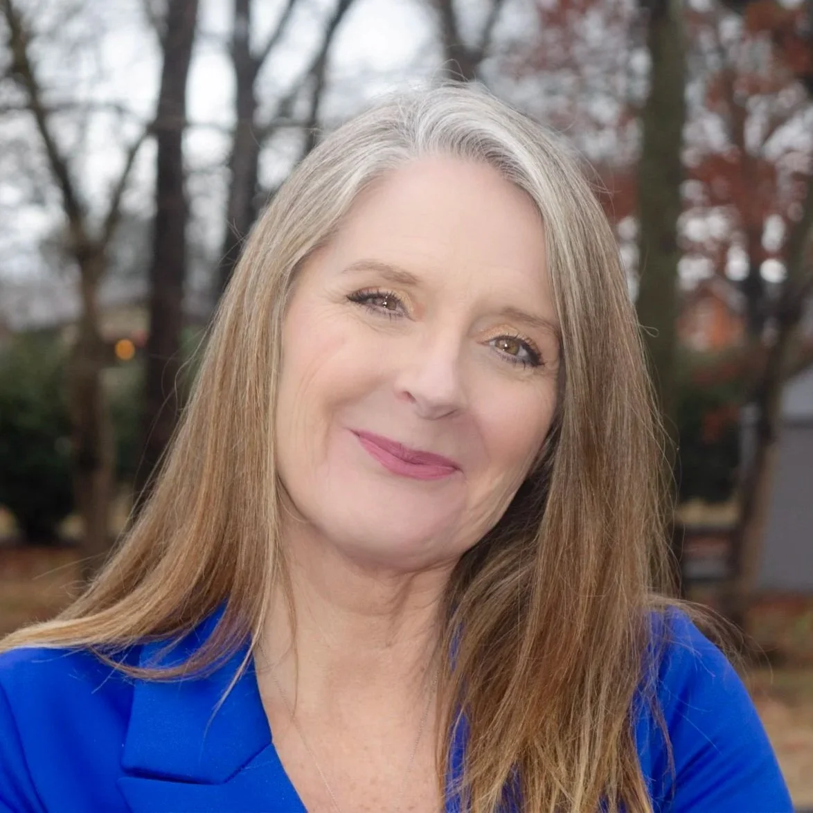A smiling woman with blonde hair wearing a blue blazer outdoors, with greenery and a brick wall in the background.