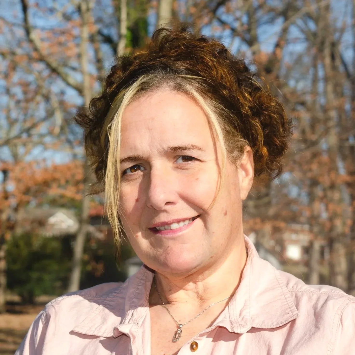 A woman with long, curly red hair smiling in front of a white textured wall.