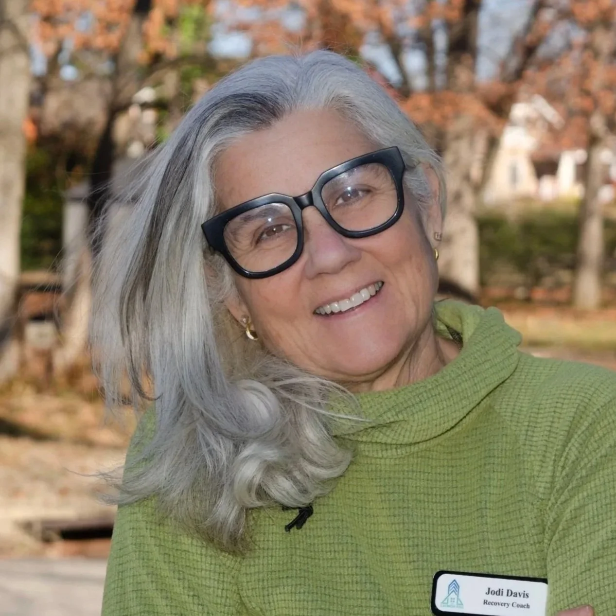 An elderly woman with gray hair in a bun, wearing glasses, a pastel scarf, and a light green top, smiling and touching her face.