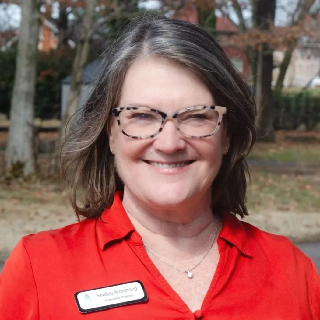 A smiling woman with short brown hair wearing a pearl headband, turquoise dress, and a necklace, speaking into a microphone.
