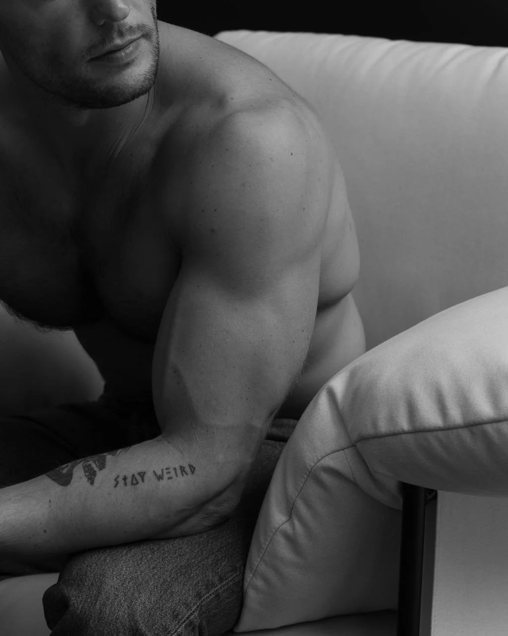 A black and white photo of a shirtless man sitting on a couch with his arm resting on his knee, showing tattoos on his arm.