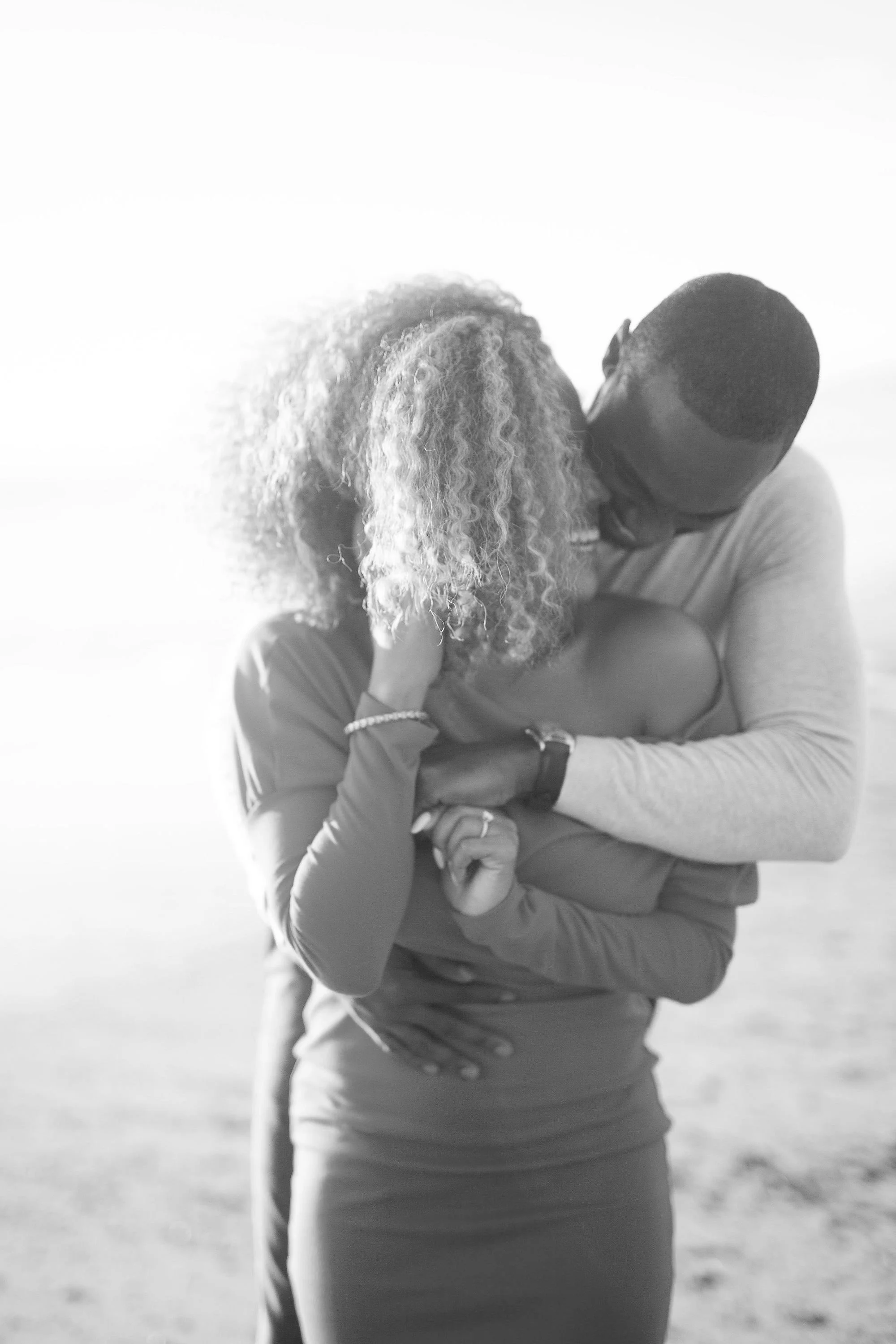 A couple kissing outdoors, with the woman wearing a long sleeve top and the man in a light-colored shirt, on a bright day.