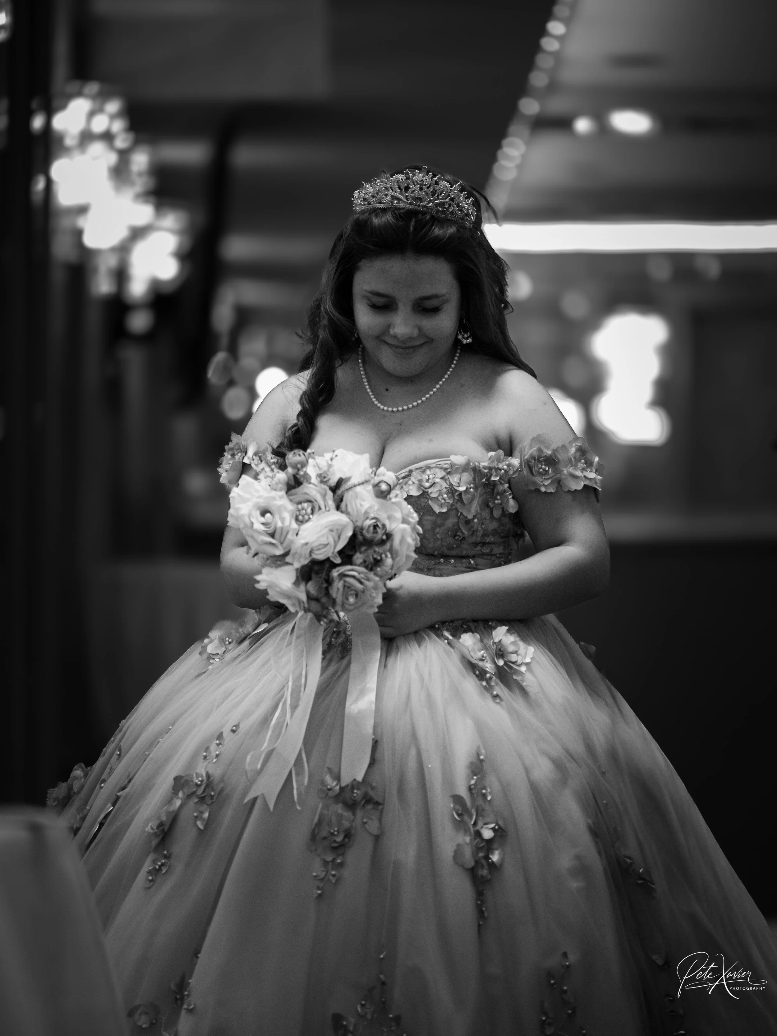 A bride in a wedding dress holding a bouquet, wearing a tiara and pearl necklace, smiling softly in an indoor setting.
