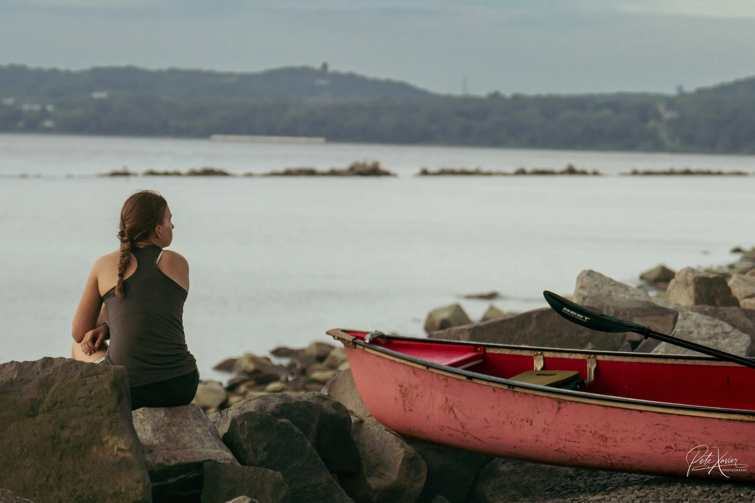 A woman with braided hair and a black tank top sitting on rocks by a body of water, with a red kayak and paddle nearby, under a cloudy sky.
