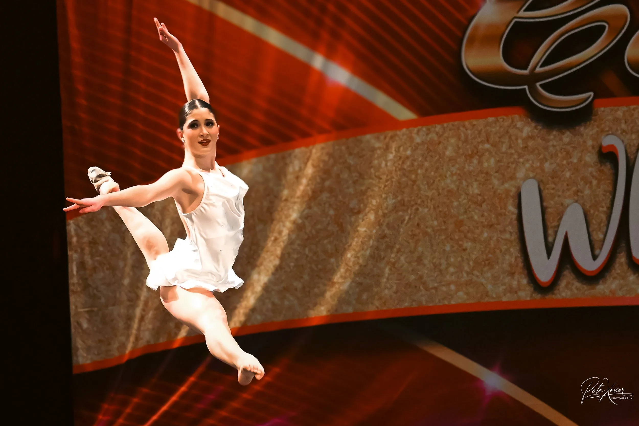 A female dancer performing a jump on stage, wearing a white dress and a big smile, with a colorful backdrop and part of a large sign visible.