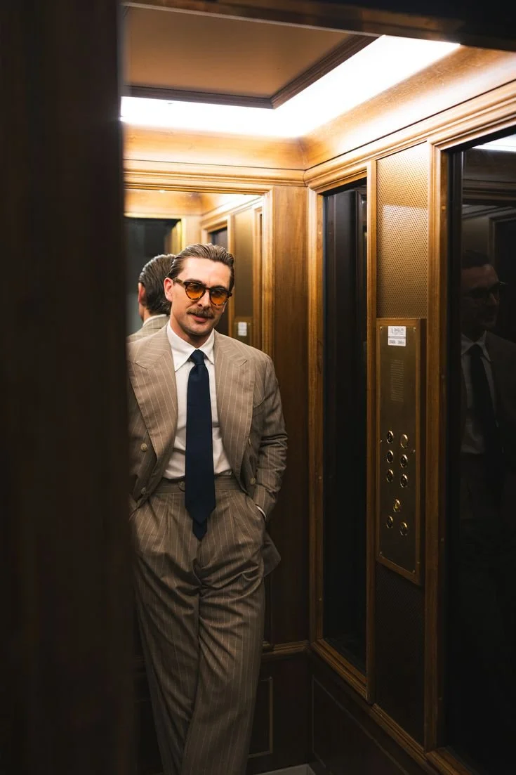 A man in a brown pinstripe suit, white shirt, and navy tie standing in an elevator with wood-paneled walls, wearing orange-tinted glasses and looking at his reflection.