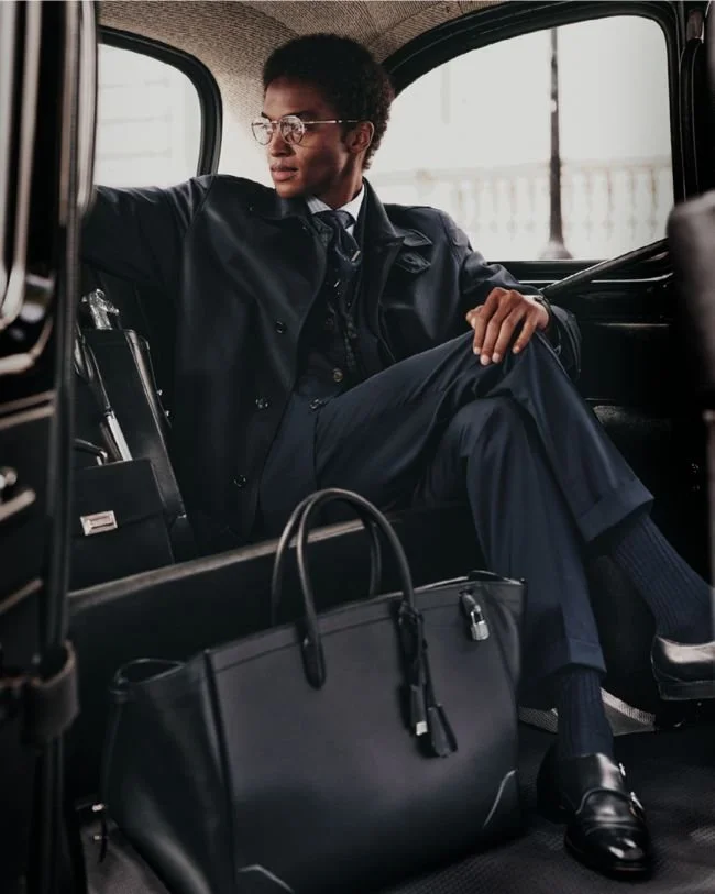 A woman in business attire sitting inside a black vehicle, with a large black handbag in front of her.