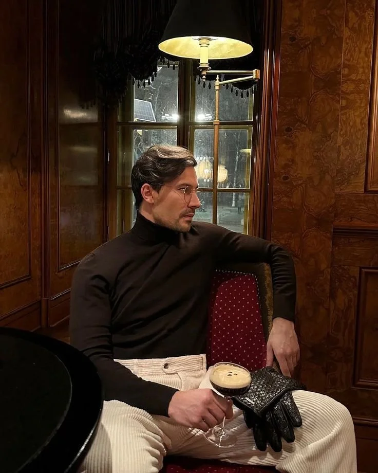 A man sitting in a vintage-style room holding a cocktail, wearing a black turtleneck and cream corduroy pants, with a pair of black gloves on his lap, near a window and a hanging lamp.