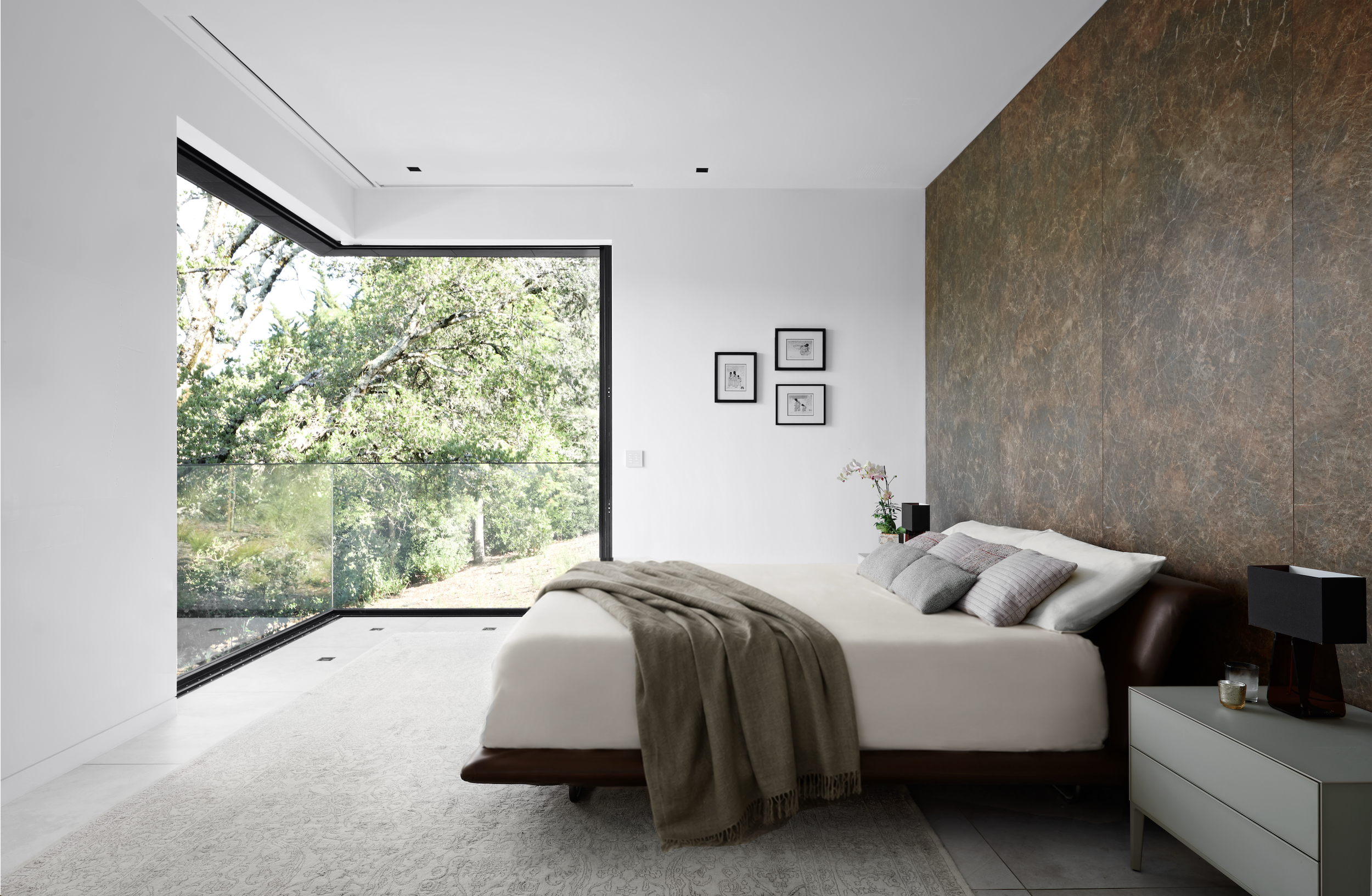 Contemporary Hillsborough bedroom featuring a dark leather platform bed, textured stone accent wall, soft neutral bedding, and floor-to-ceiling corner windows overlooking mature trees.