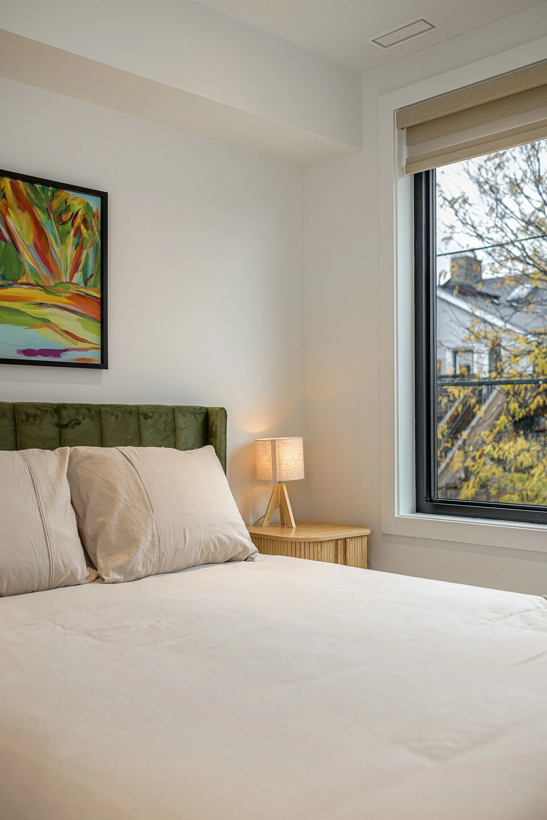 A neatly made bed with cream-colored bedding and pillows, a small wooden nightstand with a lamp, a window showing trees outside, and a colorful abstract painting on the wall.