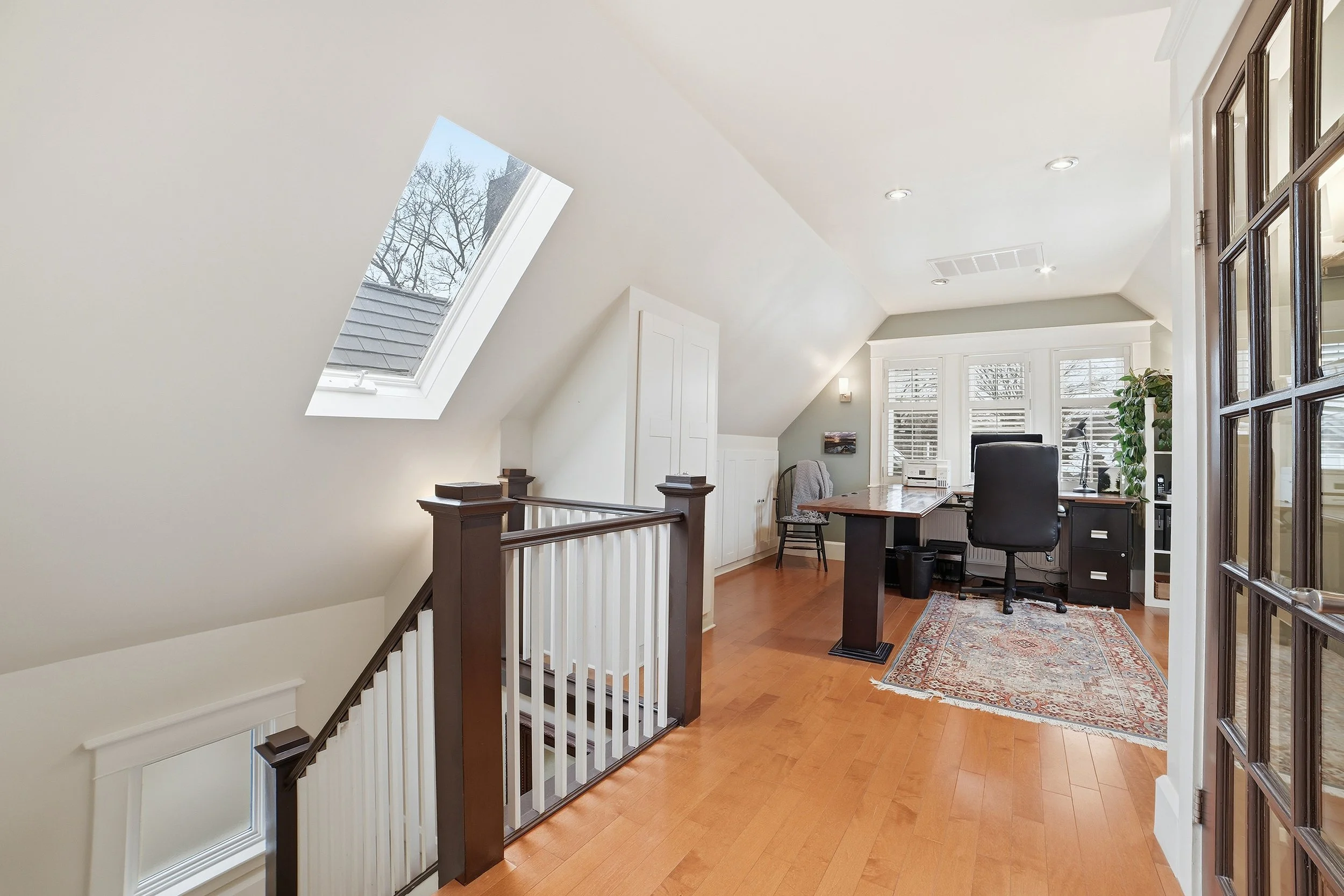 Home office space with desk, chair, and window blinds, under a sloped ceiling with a skylight and hardwood floor.