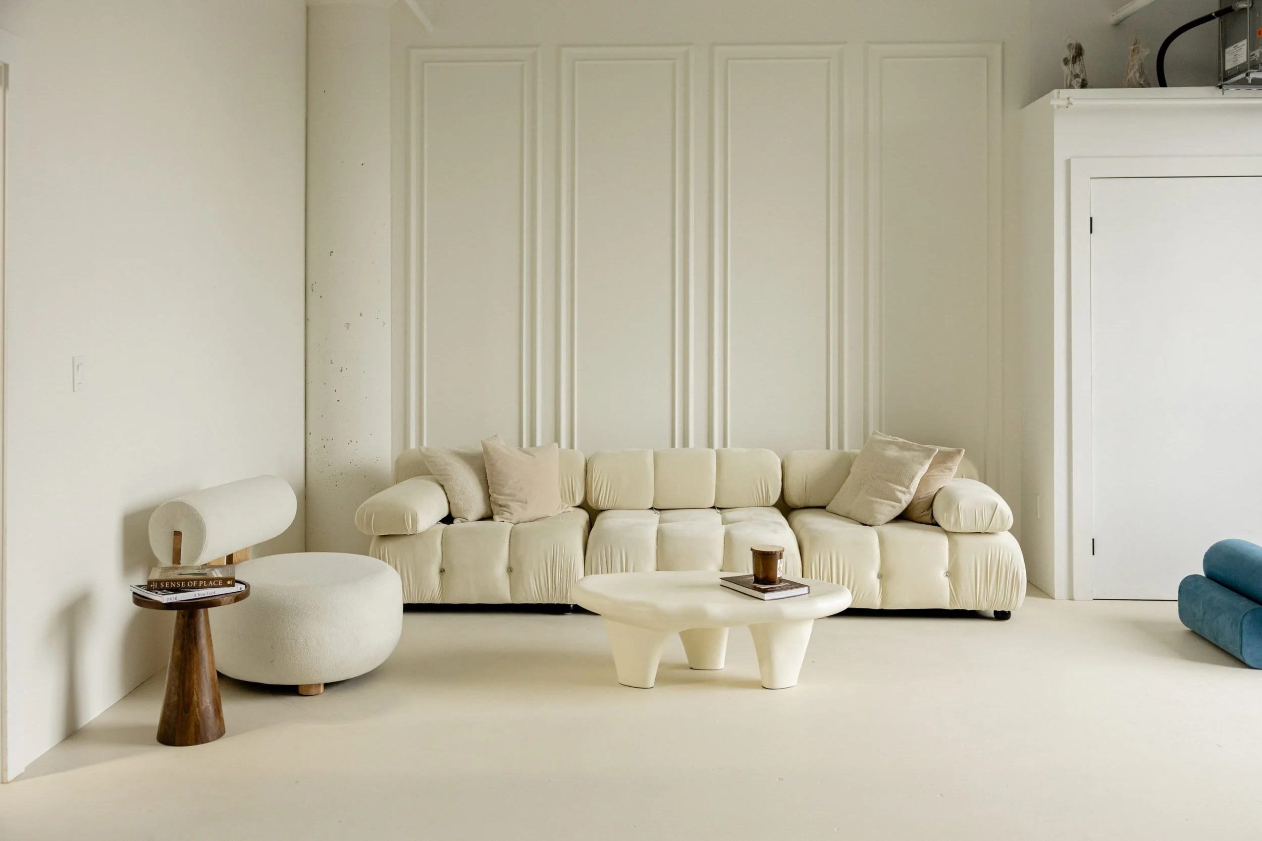 Minimalist living room with white sofa, beige and cream pillows, white side table, wooden-smoked coffee table with a candle and magazine, white armchair with a wooden side table, and part of a blue chair on the right, all set against white walls and floors.