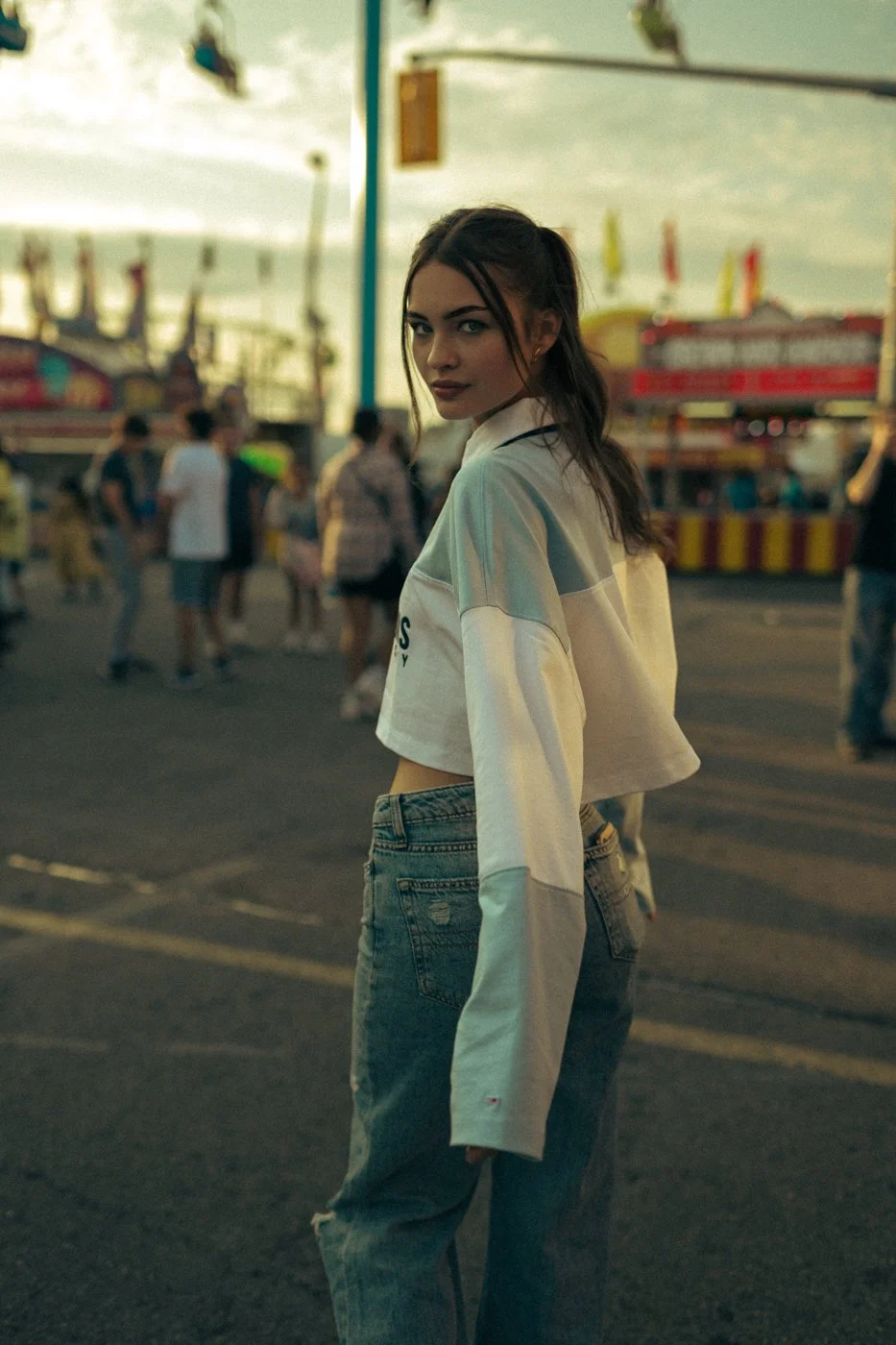 A young woman with brown hair tied in a ponytail and wearing a cropped, white and gray shirt with long sleeves, standing at a fairground with rides and booths in the background during what appears to be late afternoon or early evening.