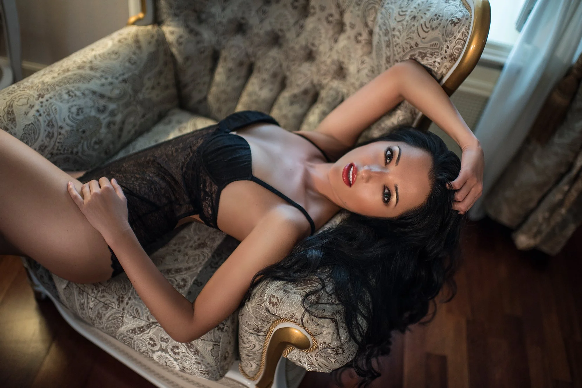 Woman in black lace lingerie reclining on an elegant armchair, arching her back to highlight natural curves in a soft-lit boudoir setting