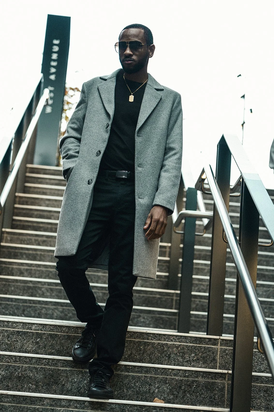 A man wearing sunglasses, a black shirt, a gray overcoat, black pants, and black shoes, standing on outdoor stairs.
