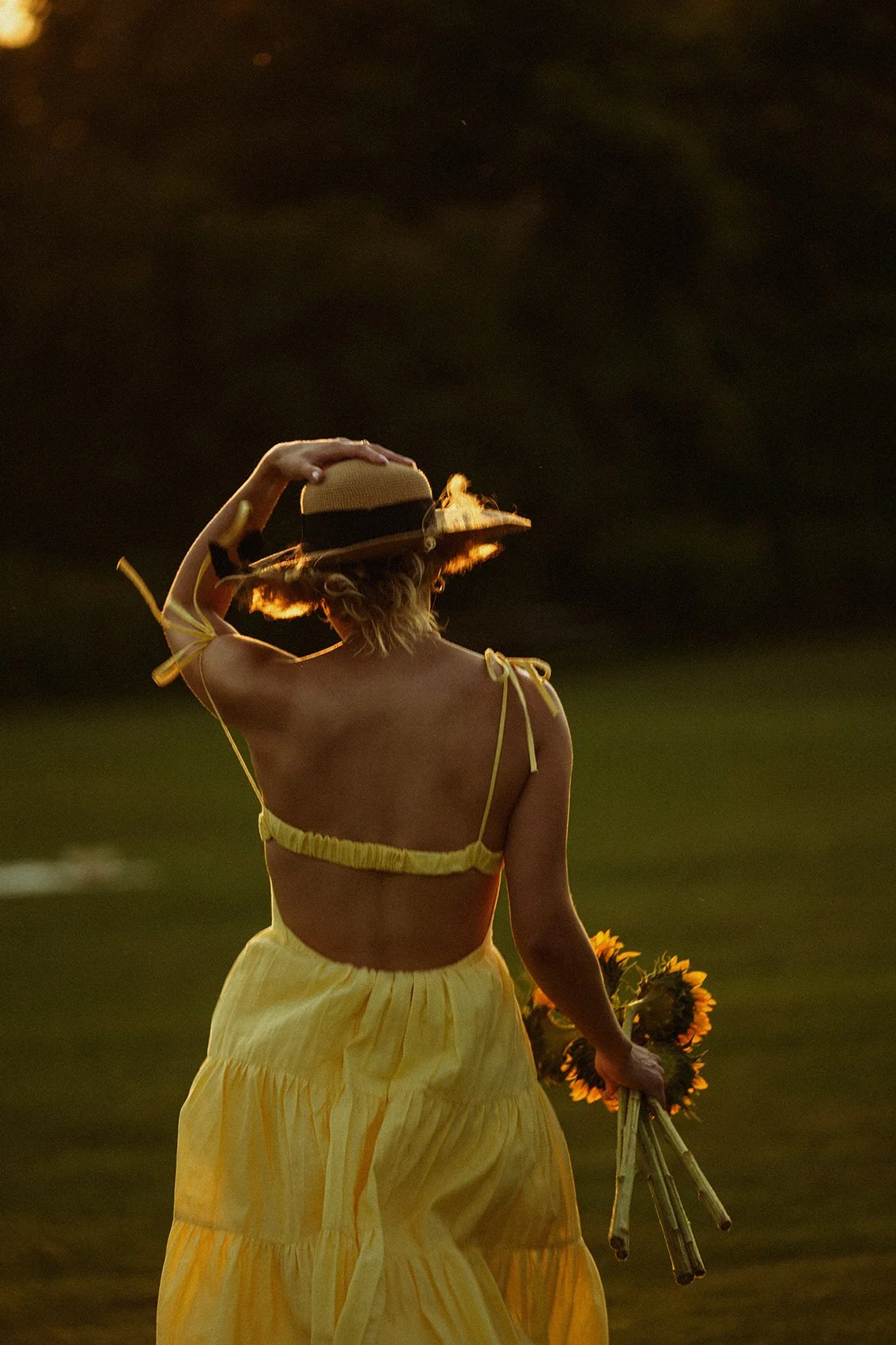 A woman in a yellow dress and straw hat holding a bouquet of sunflowers, walking outdoors at sunset.