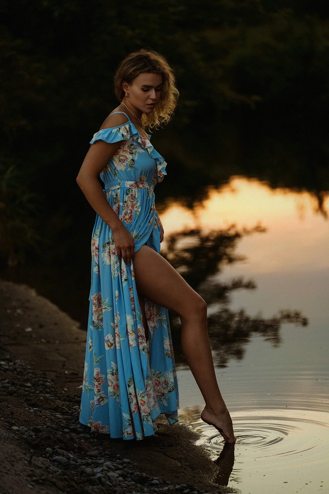 Woman in a blue floral dress touching the water at sunset by a lakeshore.
