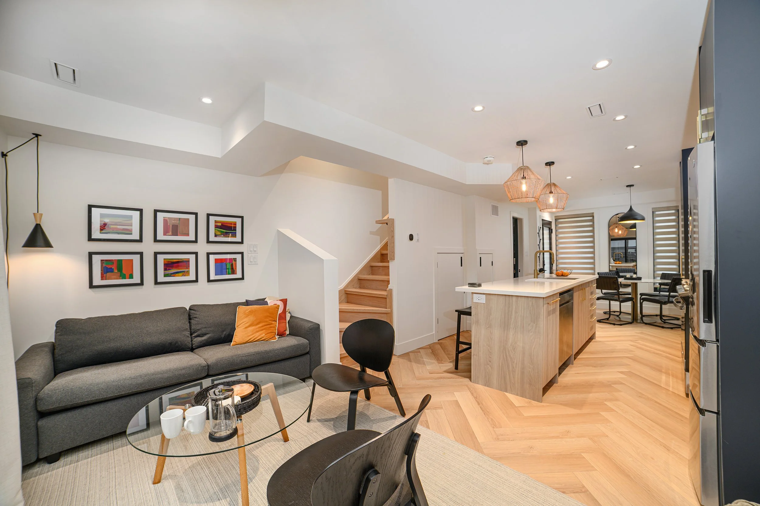 Modern open-concept living room and kitchen with gray sofa, black chairs, wooden flooring, artwork on the wall, and contemporary pendant lights.