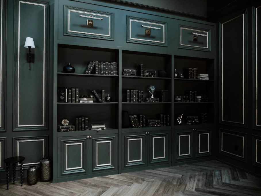 Dark green bookshelf with closed cabinets on top and bottom, filled with black and metallic decor items, against matching dark green paneled walls with black and white accents, wood floor.