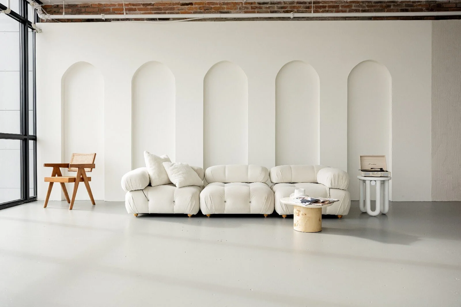 Minimalist living room with white couch, wooden chair, and vintage record player against a white wall with arched niches, large window on the left, and industrial ceiling
