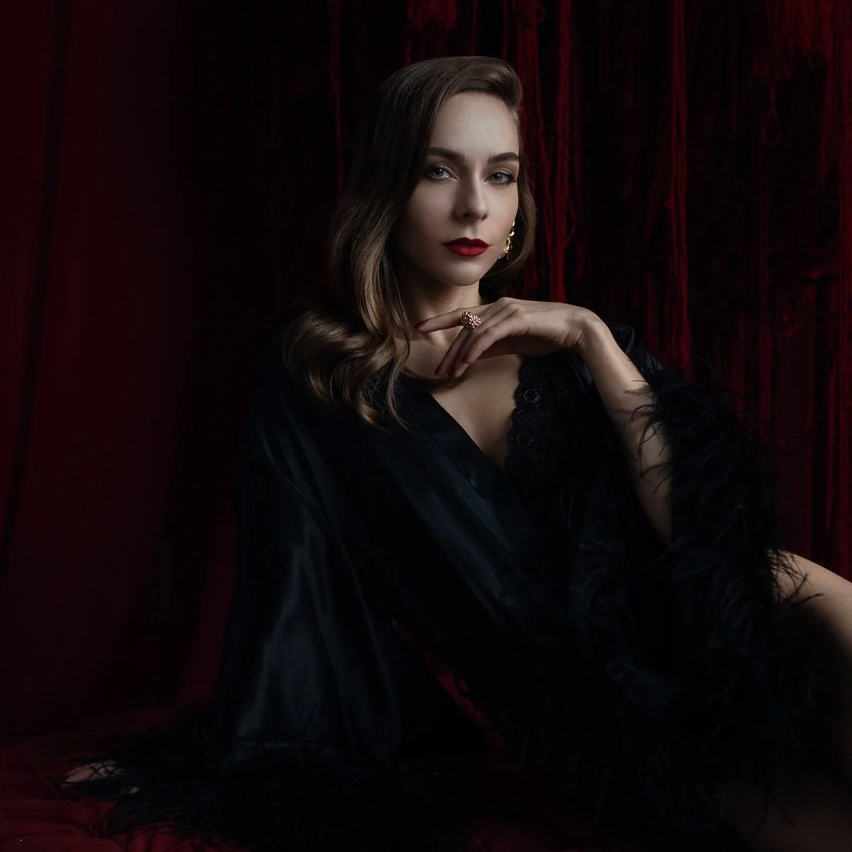 Elegant woman in black satin and feather-trimmed robe reclining on a red velvet couch, showcasing confident boudoir posing with moody lighting