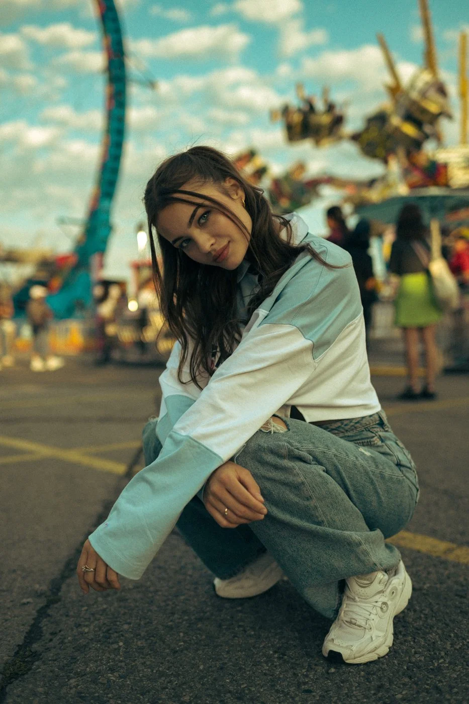 A young woman with long dark hair, dressed in a light-colored jacket, baggy jeans, and white sneakers, squatting on the pavement at an amusement park with a carnival ride and colorful sky in the background.