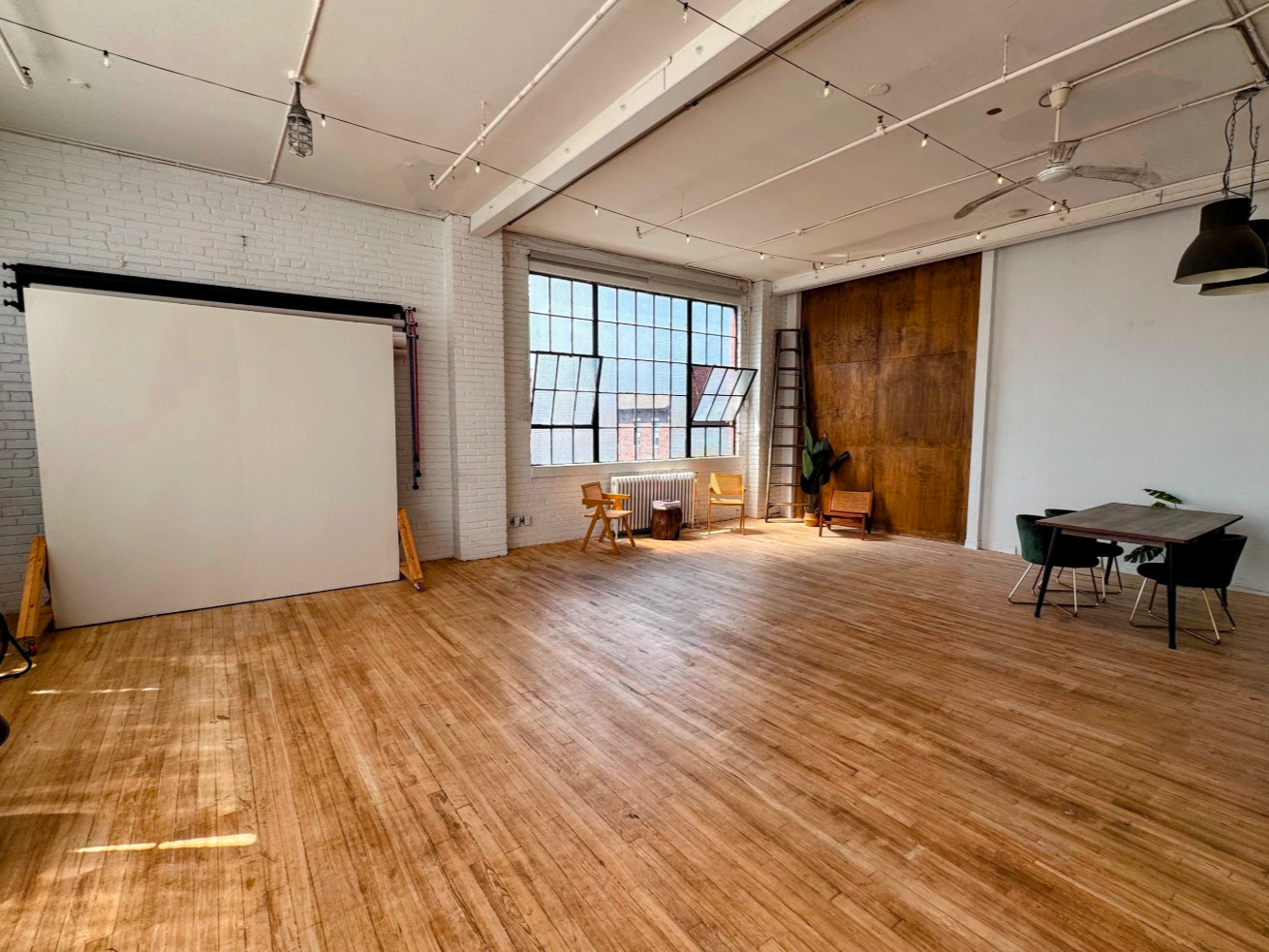 Spacious room with hardwood floors, large industrial-style windows, white brick walls, a white backdrop, a ladder, a small table with chairs, and a corner with plants.
