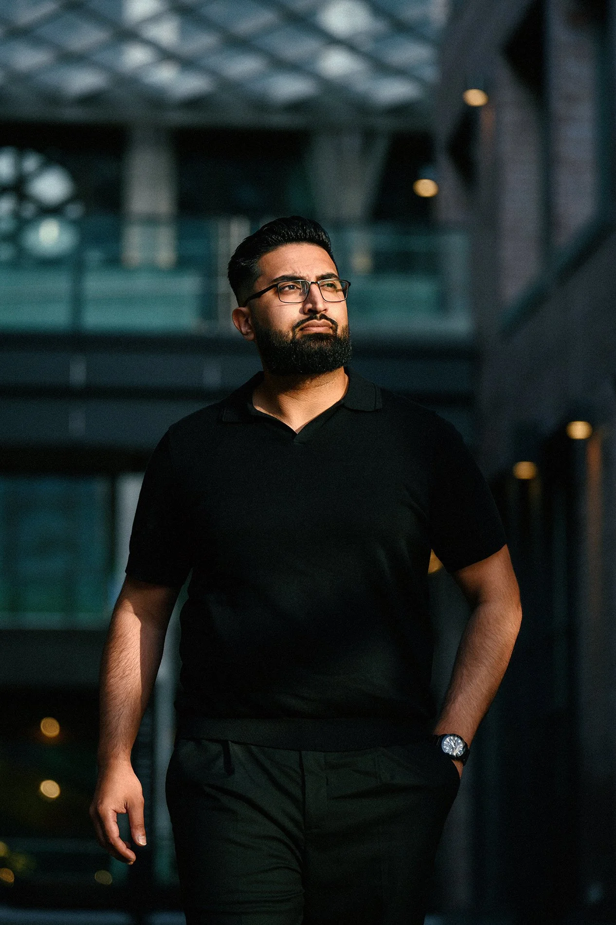 A man with dark hair, beard, glasses, wearing a black polo shirt and black pants, walking outdoors at dusk with modern buildings in the background.