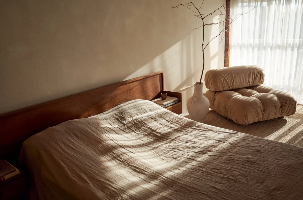 A bedroom with sunlight casting shadows on a bed with a beige cover, a wooden headboard, a side table with books and a candle, a large beige cushioned chair, and a tall vase with a bare tree branch, near a window with sheer curtains.