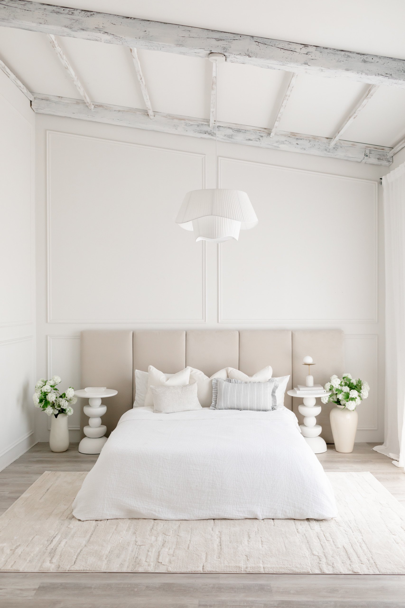 A minimalist bedroom with a white bed, beige headboard, two white nightstands with vases of white flowers, a modern white pendant light, and white walls with decorative paneling.