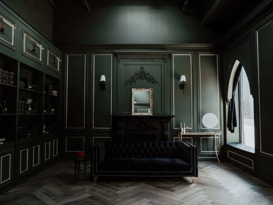 A dark green living room with black ornate furniture, a tufted black sofa, gold-framed mirror, wall sconces, a window seat, and decorative shelving.
