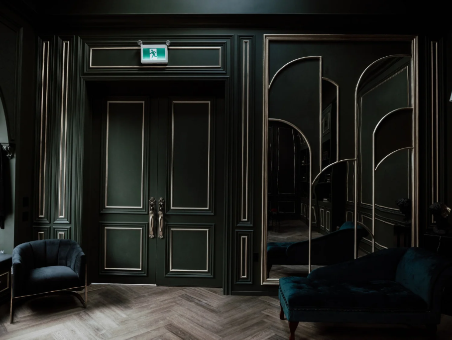 Dark green double doors with silver handles in an elegant room with dark green paneling, a mirror, a black armchair, and a teal sofa.