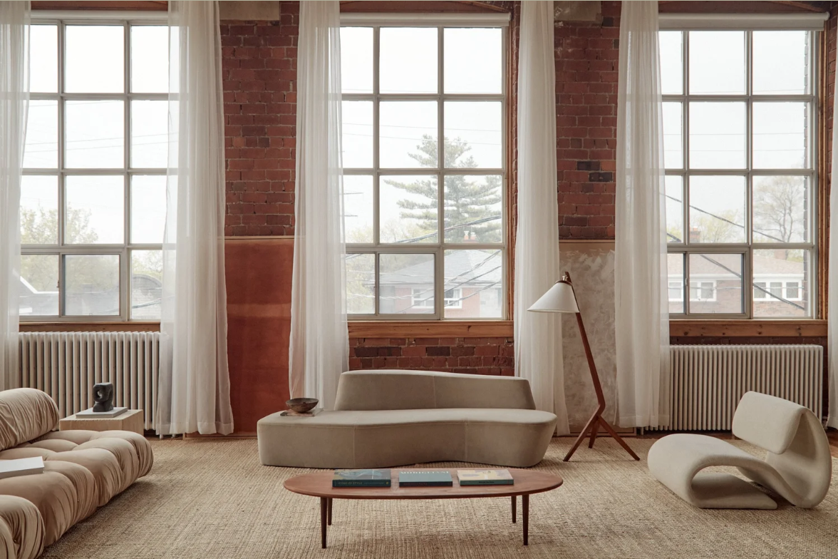 Living room with large windows, sheer white curtains, brick wall, beige sofa, modern white lounge chair, wooden coffee table with magazines, floor lamp, and radiator.