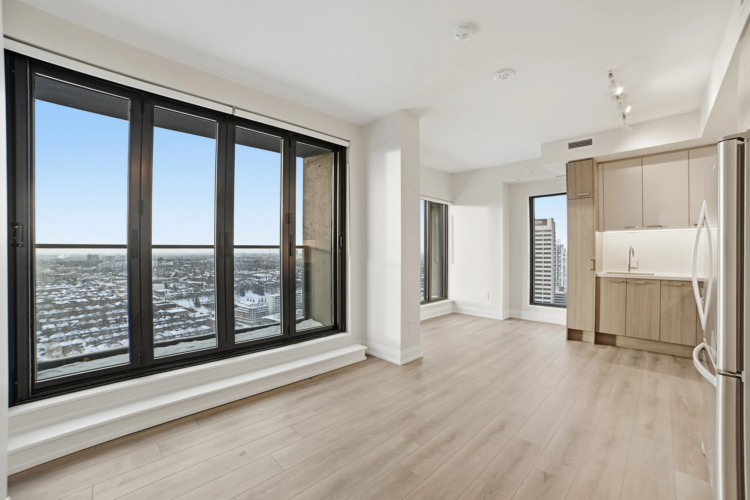 Empty modern apartment with large windows showing city view, light wood flooring, and minimalist kitchen with beige cabinets and stainless steel refrigerator.