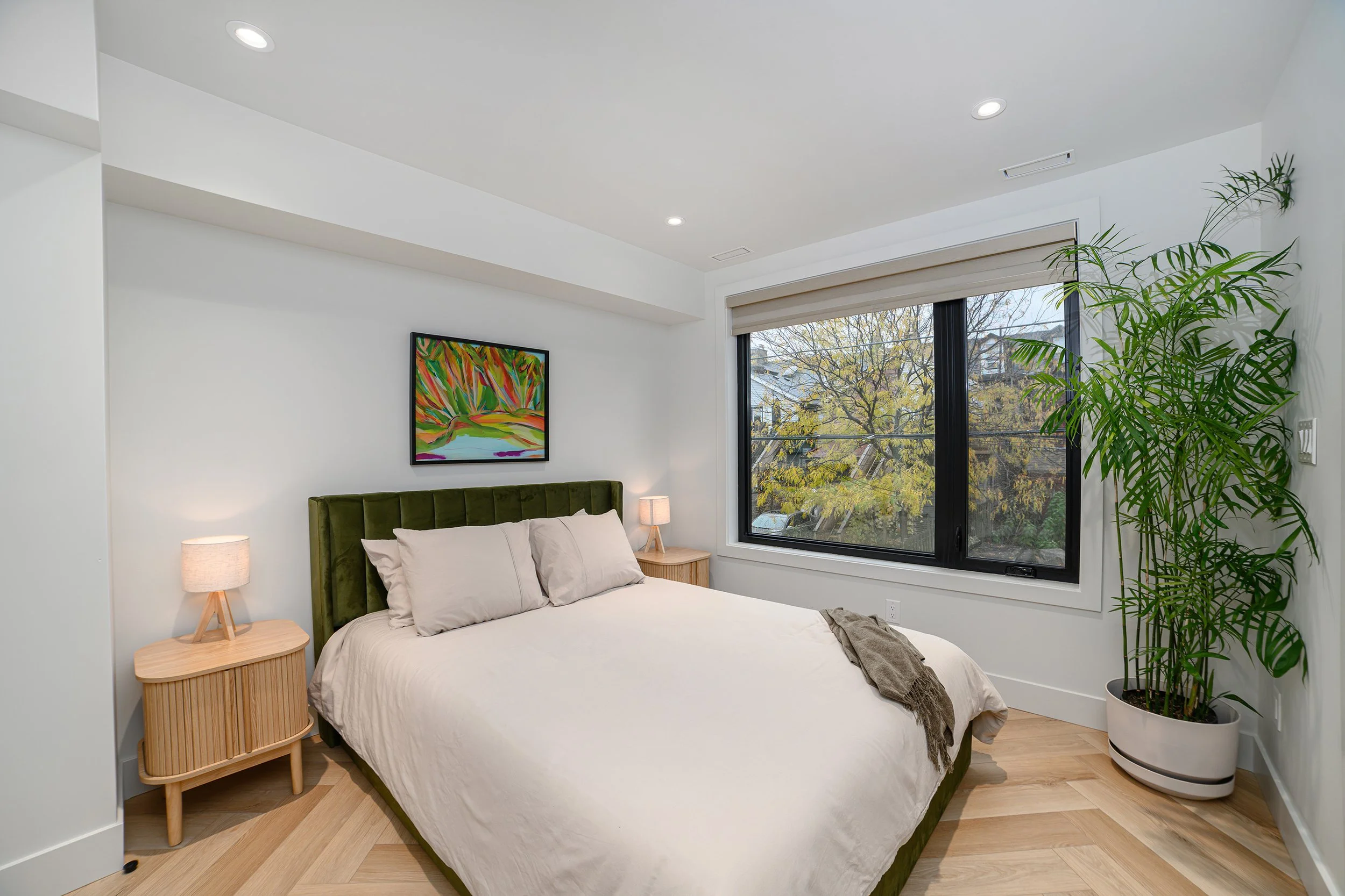 Bedroom with a green upholstered headboard, white bedding, two wooden nightstands with lamps, a large window with a view of trees, and a potted plant in the corner.