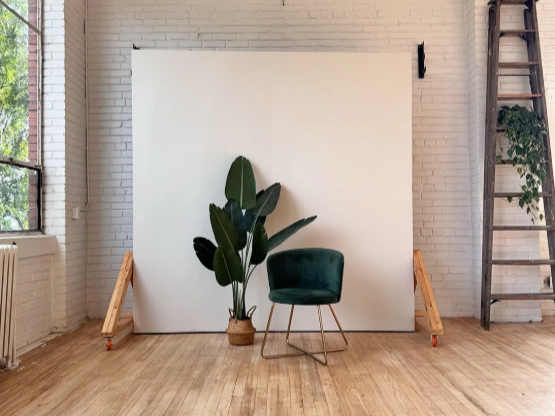 A photography studio setup with a white backdrop, a houseplant in a basket, a dark green chair, and a wooden ladder against a brick wall.