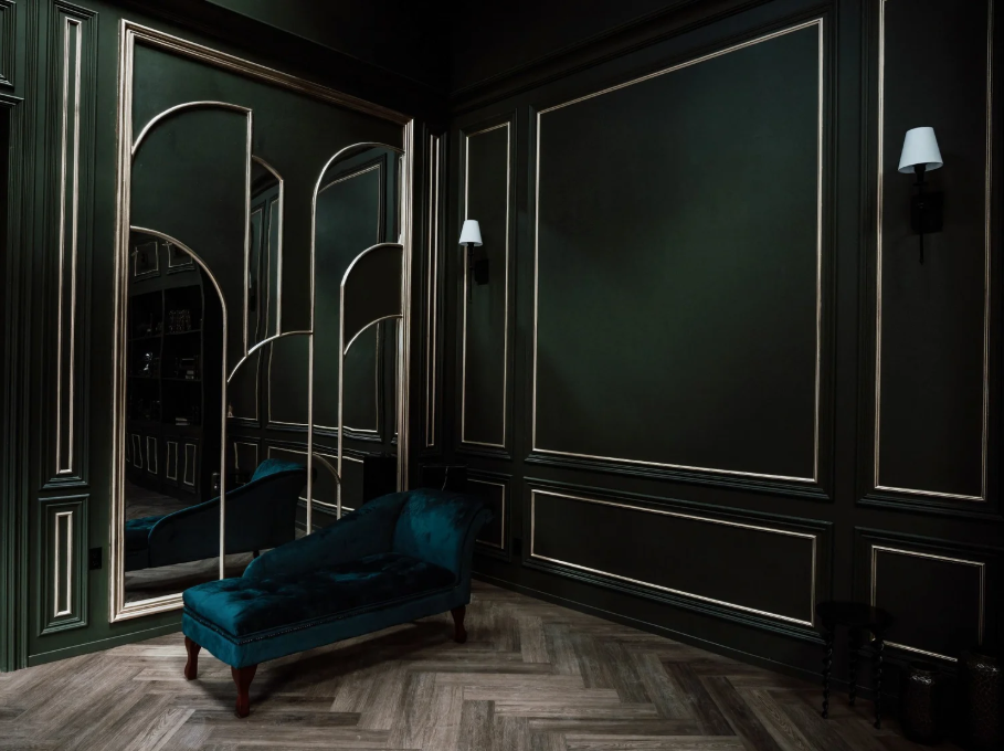 Luxurious dark green room with gold accents, decorative molding, a velvet chaise lounge, a large mirror, and wall sconces.